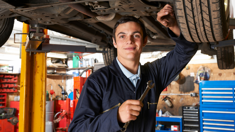 Car mechanic apprentice stood under a car holding a spanner and smiling for the camera