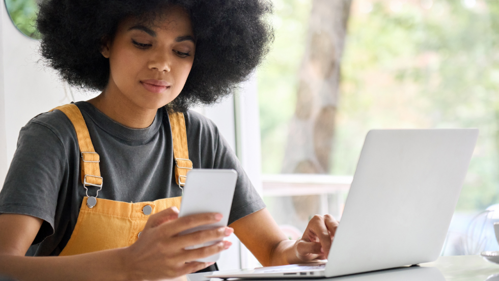 Young person sat at a table with a laptop. They are also reading their phone