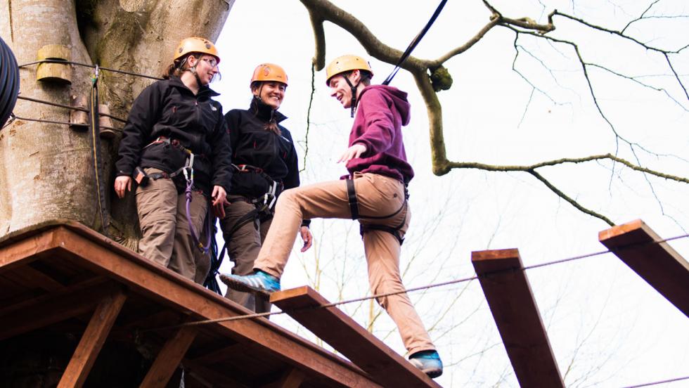 A person steps wearing climbing safety gear steps onto a wooden platform from a bridge while two others look on