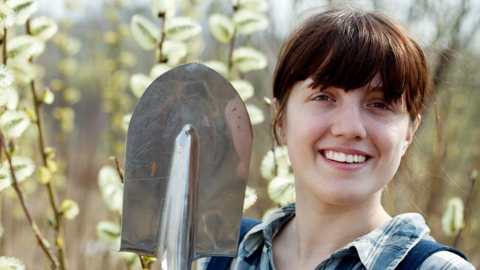 A young woman in a checked shirt stands in a field with a shovel