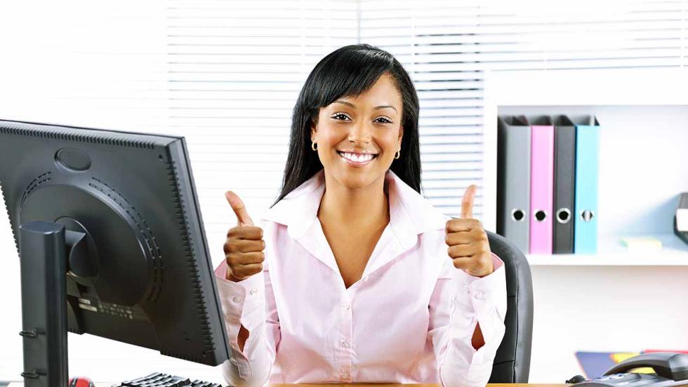 A young woman sits behind a desk with a computer, grinning and giving two tumbs up