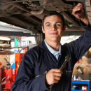 Car mechanic apprentice stood under a car holding a spanner and smiling for the camera