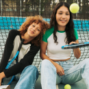 Two friends sat down on a tennis court. One is bouncing a tennis ball on a tennis racket.