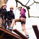 A person steps wearing climbing safety gear steps onto a wooden platform from a bridge while two others look on